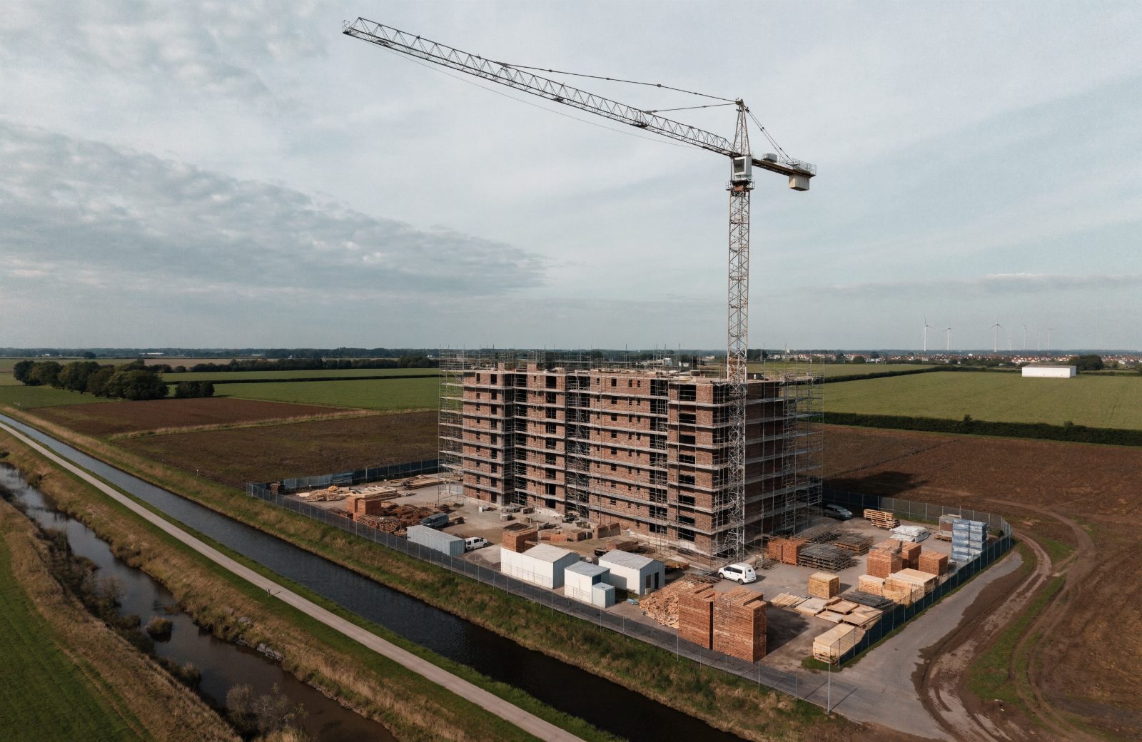 Bouwplaats van bovenaf gezien, met een half opgetrokken bakstenen gebouw in de steigers, een torenkraan, bouwketen en een afgezet terrein in een Nederlands landschap