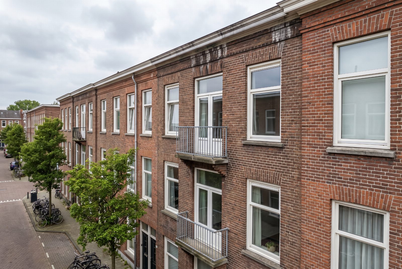 Bakstenen gevel van een Nederlands appartementengebouw met witte kozijnen en een balkon, gezien vanuit de lucht op verdiepingshoogte